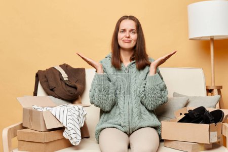 Photo for Sad upset woman sitting on sofa among boxes with clothing against beige wall shrugging shoulders looking at camera with unhappy expression unpacking things. - Royalty Free Image