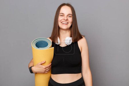 Photo for Sport for heath. Smiling woman wearing black sportswear holding yoga mat standing isolated over gray background looking at camera with satisfied face after yoga training. - Royalty Free Image