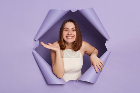 Photo for Cute smiling happy young brunette woman in casual clothing breaking through purple paper hole looking at camera with positive facial expression. - Royalty Free Image