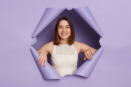Photo for Charming smiling young brunette woman in casual clothing breaking through purple paper hole being in good mood looking with happy face friendly expression. - Royalty Free Image