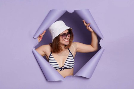 Photo for Friendly attractive woman wearing swimwear panama and sunglasses breaking through purple paper background looking away with happy face expressing positive emotions. - Royalty Free Image