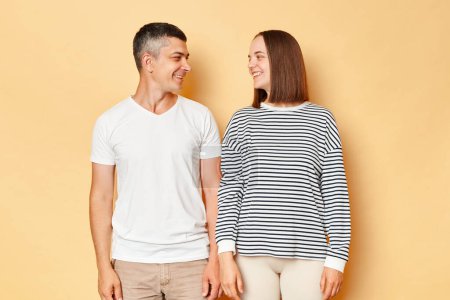 Photo for Pretty happy loving young couple wife and husband wearing casual attires standing isolated over beige background looking at each other with smiles and gentles, expressing love. - Royalty Free Image