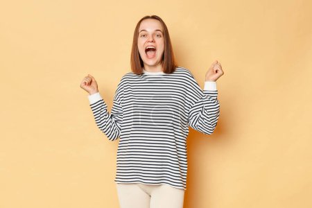 Photo for Extremely happy cheerful brown haired woman wearing striped shirt standing isolated over beige background rejoicing hearing good news celebrating victory clenched fists. - Royalty Free Image