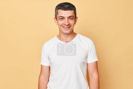 Photo for Smiling handsome man wearing white casual t-shirt standing isolated over beige background looking at camera being in good mood expressing positive emotions. - Royalty Free Image