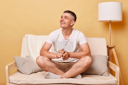 Photo for Satisfied dreamy man wearing white T-shirt and shorts sitting on sofa against beige background looking away with toothy smile expressing positive emotions. - Royalty Free Image