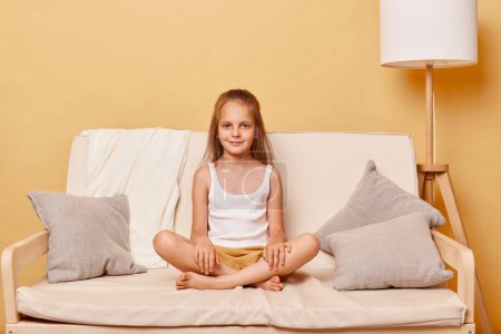 Photo for Cute charming little girl wearing casual clothing sitting on sofa at home against beige wall resting alone looking at camera with calm facial expression. - Royalty Free Image