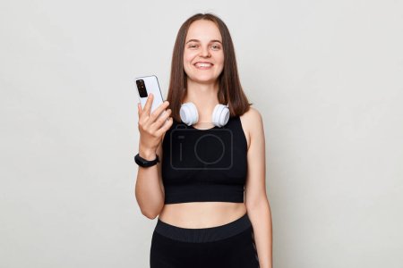 Photo for Smiling cheerful slim woman with headphones wearing sportswear posing against gray background using mobile phone sporty app. - Royalty Free Image