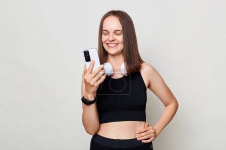 Photo for Attractive joyful slim woman with headphones wearing sportswear posing against gray background using cell phoner after workout expressing positive emotions. - Royalty Free Image