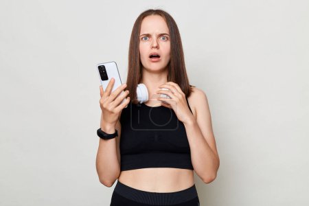 Photo for Shocked slim woman with headphones wearing sportswear posing against gray background reading new in social network berowsing internet. - Royalty Free Image