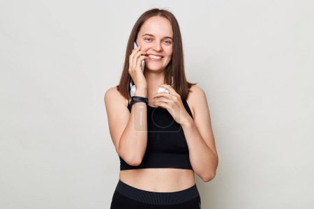 Photo for Cheerful optimistic slim woman with headphones wearing sportswear posing against gray background talking on cell phone, calling to friends, mobile call. - Royalty Free Image