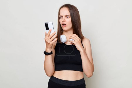 Photo for Attractive shocked surprised mslim woman with headphones wearing sportswear posing against gray background holding mobile phone looking at display with astonished face. - Royalty Free Image