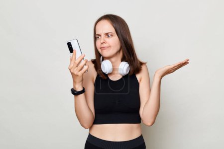 Photo for Puzzled confused slim woman with headphones wearing sportswear posing against gray background using mobile phone having problems with app. - Royalty Free Image
