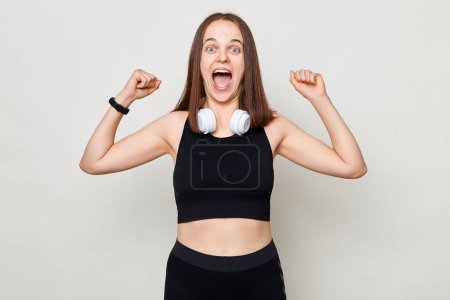 Photo for Excited amazed slim woman with headphones wearing sportswear posing against gray background standing raised her arms showing her power screaming. - Royalty Free Image