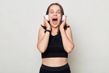 Photo for Happy excited slim woman with headphones wearing sportswear posing against gray background listening music in headphones screaming with excitement. - Royalty Free Image