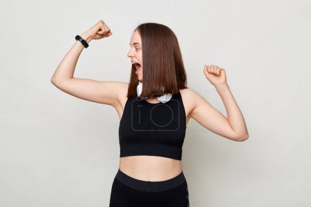Photo for Extremely happy slim woman with headphones wearing sportswear posing against gray background looking at her biceps with amazed excited expression. - Royalty Free Image