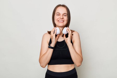 Photo for Smiling positive cheerful slim woman with headphones wearing sportswear posing against gray background lookign at camera with positive expression. - Royalty Free Image