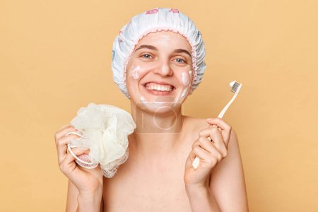 Foto de Atractiva mujer sonriente con gorra de ducha de pie aislada sobre fondo beige sosteniendo el cepillo de dientes posando después de tomar la ducha. - Imagen libre de derechos