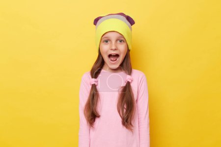 Photo for Extremely happy amazed little girl with ponytails wearing beanie hat standing isolated over yellow background screaming with widely open mouth. - Royalty Free Image