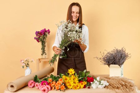 Photo for Plant store. Floral bouquet. Flower shop. Cheerful joyful young woman florist wearing brown apron posing at her workplace standing isolated over beige background making beautiful bouquet. - Royalty Free Image