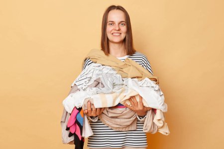 Foto de Sonriente alegre mujer morena positiva sosteniendo pila de ropa en manos aisladas sobre fondo beige haciendo tareas domésticas ordenando su armario. - Imagen libre de derechos