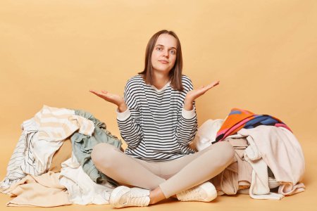 Photo for Uncertain confused Caucasian woman posing near heap of multicolored unsorted clothes isolated over beige background needs decluttering sorting her attires sitting with crossed legs - Royalty Free Image