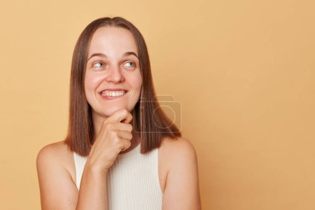 Photo for Dreaming cheerful brown haired young woman wearing casual clothing standing isolated over beige background holding her chin looking way at copy space for advertisement. - Royalty Free Image