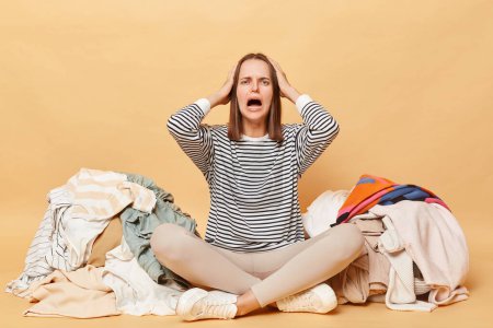 Photo for Despair woman surrounded by clothing for recycling isolated over beige background sitting on floor with colorful unfolded clothes busy doing wardrobe cleaning. - Royalty Free Image