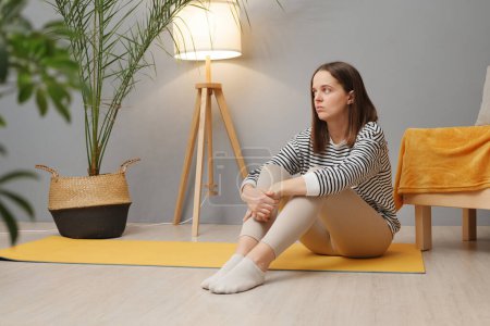 Photo for Young adult depressed brown haired attractive woman sitting on fitness mat after training at home wearing casual clothing looking away with pensive expression - Royalty Free Image