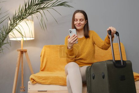 Téléchargez les photos : Jolie femme d'affaires caucasienne portant un pull orange assis avec des bagages sur le canapé dans la chambre d'hôtel attendant son départ naviguant sur Internet via smartphone - en image libre de droit