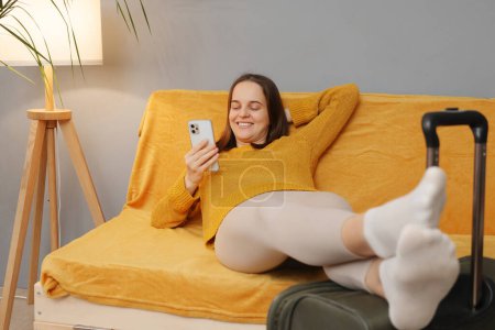 Téléchargez les photos : Belle femme blanche joyeuse portant un pull orange assis avec des bagages sur le canapé en regardant l'écran de téléphone portable avec sourire naviguer sur Internet avant de quitter l'hôtel - en image libre de droit