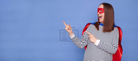 Joyful happy overjoyed Caucasian brown haired woman wearing superhero costume and striped shirt isolated over blue background indicating at advertising area excellent promotion