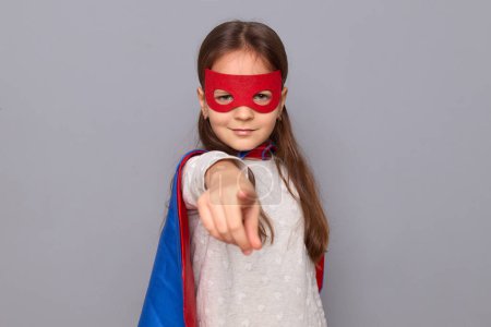 Confident little girl wearing superhero costume and mask isolated over gray background pointing finger to camera choosing you looking with strict expression