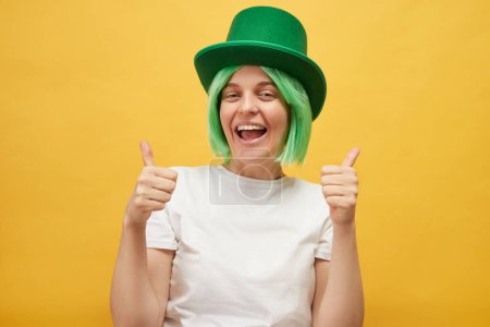 Genießen für St. Patrick 's Day Feiern. Entzückte kaukasische Frau mit grünem Koboldhut steht isoliert über gelbem Hintergrund und zeigt Daumen hoch wie eine Geste