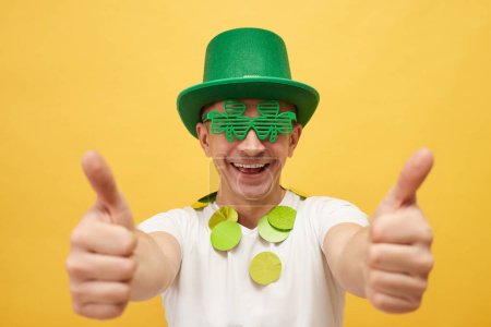 Nationaler Urlaubsspaß. Lächelnder Mann mit festlich grünem Hut und Shamrock-Brille steht isoliert vor gelbem Hintergrund und zeigt Daumen hoch wie eine Geste