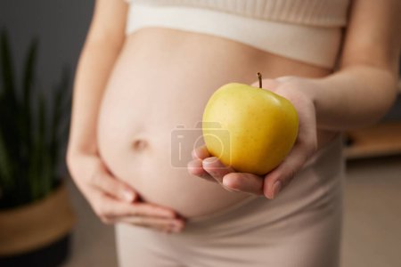 Unknown beautiful pregnant woman with bare belly standing in home interior holding fresh organic yellow apple prefers healthy eating while expecting baby