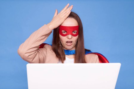 Upset sad woman wearing superhero costume standing isolated over blue background working on laptop having problems with work showing facepalm gesture