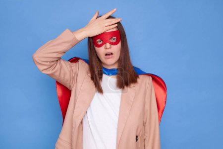 Displeased disappointed woman wearing superhero costume standing isolated over blue background having troubles showing facepalm gesture forgot something pleasant