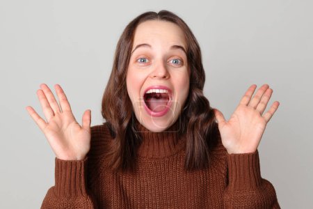 Extremely happy Caucasian woman wearing brown sweater standing isolated over light gray background raising her arms screaming with positive emotions rejoicing