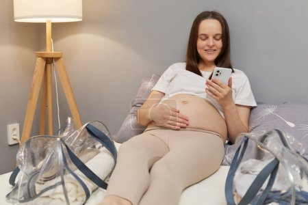 Schöne entspannte Schwangere, die auf dem Bett liegt und ihren nackten Bauch berührt, indem sie mit dem Smartphone im Internet surft und Informationen über die Geburt liest