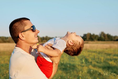 Nahaufnahme eines jungen Vaters mit seiner kleinen Tochter, die Spaß im grünen Gras hat und sich umarmt und die glückliche gemeinsame Zeit in freier Natur genießt