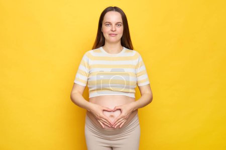Mujer embarazada caucásica hermosa adulta de cabello castaño con vientre desnudo usando top casual aislado sobre fondo amarillo tocando su vientre mostrando símbolo de amor haciendo con las manos