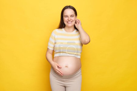 Schöne glücklich entzückte kaukasische Schwangere mit nacktem Bauch trägt lässiges Oberteil isoliert über gelbem Hintergrund und schaut mit zufriedenem Gesicht in die Kamera