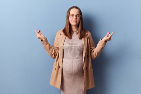Calm relaxed pregnant elegant woman in beautiful dress and jacket posing isolated over blue background standing with mudra gesture meditating trying to calm down