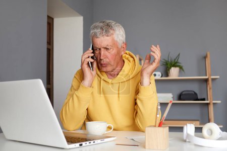 Gray haired old man wearing yellow hoodie having phone conversation sitting at table in his home workplace speaking with his colleague having work discussion