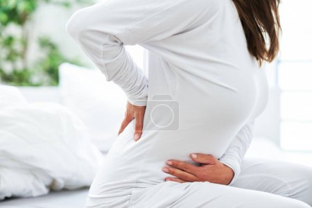 Téléchargez les photos : Jeune femme brune essuie son problème de dos dans le lit. Photo de haute qualité - en image libre de droit