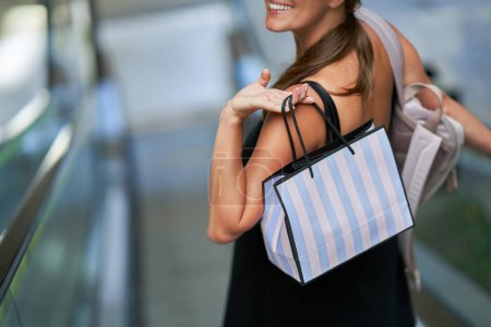 Une jeune femme sourit joyeusement en portant un sac à provisions sur l'escalier roulant dans un centre commercial branché