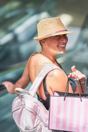 Una mujer joven sonríe felizmente mientras lleva una bolsa de compras en la escalera mecánica en un centro comercial de moda