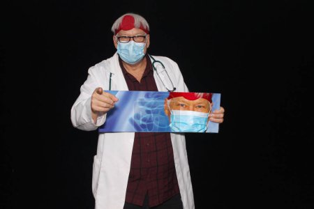 Foto de Médico. Un médico amable y amable sonríe mientras está de pie con un cartel con su retrato en él con un fondo negro. Médico masculino caucásico sonriente con uniforme médico blanco y estetoscopio - Imagen libre de derechos