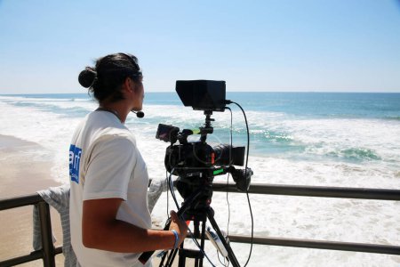 Téléchargez les photos : Huntington Beach, Californie. - USA - 19 septembre 2022 : Opérateur de caméra de presse. Un homme utilise sa caméra de nouvelles pour enregistrer et transmettre la compétition de surf à Huntington Beach en Californie. Les surfeurs rivalisent. - en image libre de droit