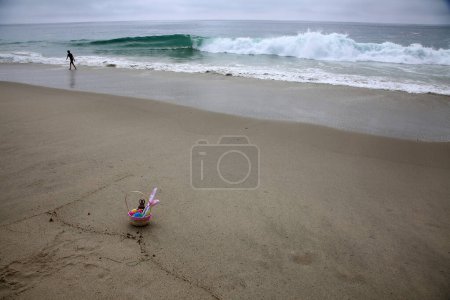 Téléchargez les photos : Panier de Pâques dans le sable près de l'océan. Un panier de Pâques avec de l'herbe verte, un lapin en chocolat, des œufs en plastique et des oreilles de lapin - en image libre de droit
